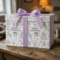 Black and white birthday-themed coloring wrapping paper featuring cakes, balloons, party hats, and stars, designed for kids gift wrap and party activity on a gift  box with purple ribbon on a wooden surface.
