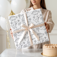 Person holding two wrapped presents with dinosaur patterns and a cake on a table.