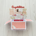Baby-themed open card with teddy bear, pram, and baby booties on a wooden background