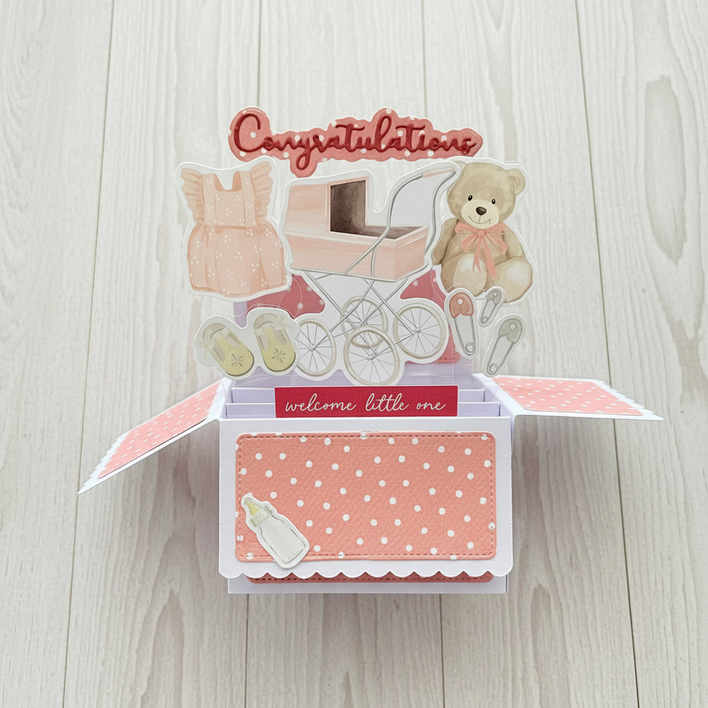 Baby-themed open card with teddy bear, pram, and baby booties on a wooden background