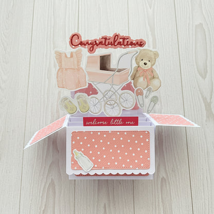 Baby-themed open card with teddy bear, pram, and baby booties on a wooden background