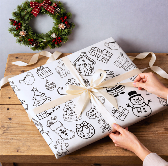 Gift wrapped in white paper with hand drawn Christmas designs suitable for coloring in, tied with a ribbon, on a wooden table with a wreath in the background.
