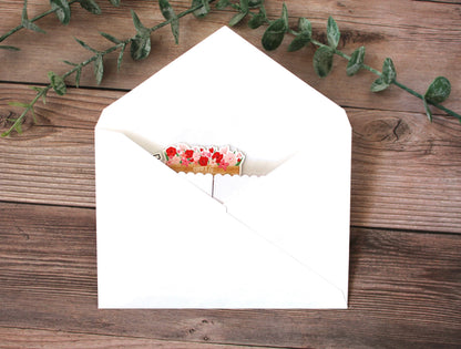 Open white envelope with a small card inside on a wooden surface with green leaves.