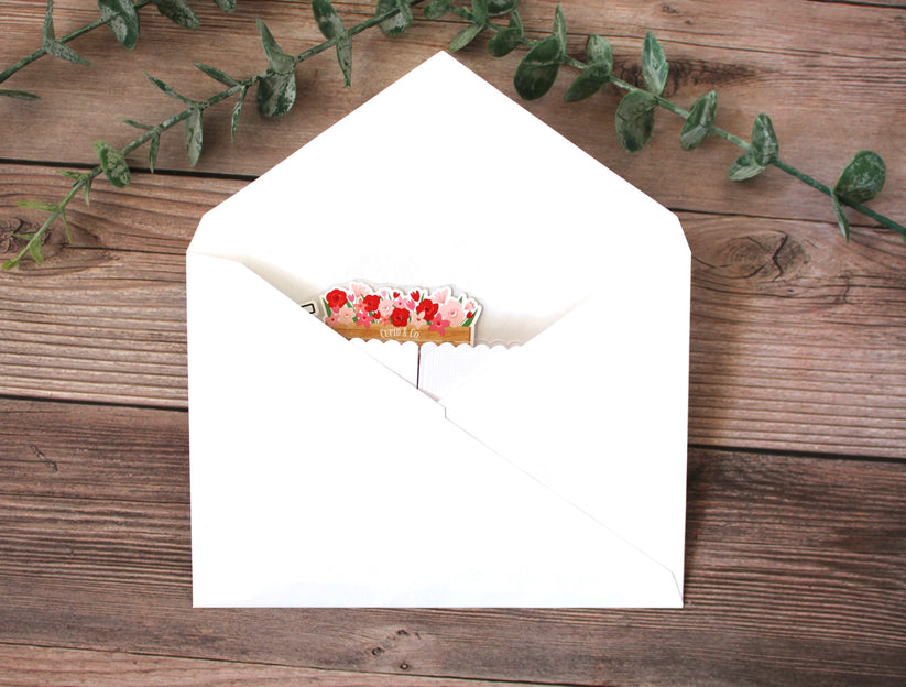 Open white envelope with a small card inside on a wooden surface with green leaves.