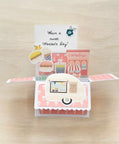 Pop-up card with coffeehouse-themed illustrations and 'Have a sweet Mother's Day!' text on a wooden surface.
