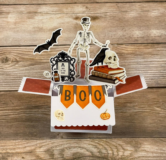 Halloween-themed pop-up card with skeleton, bat, and skull on a wooden surface