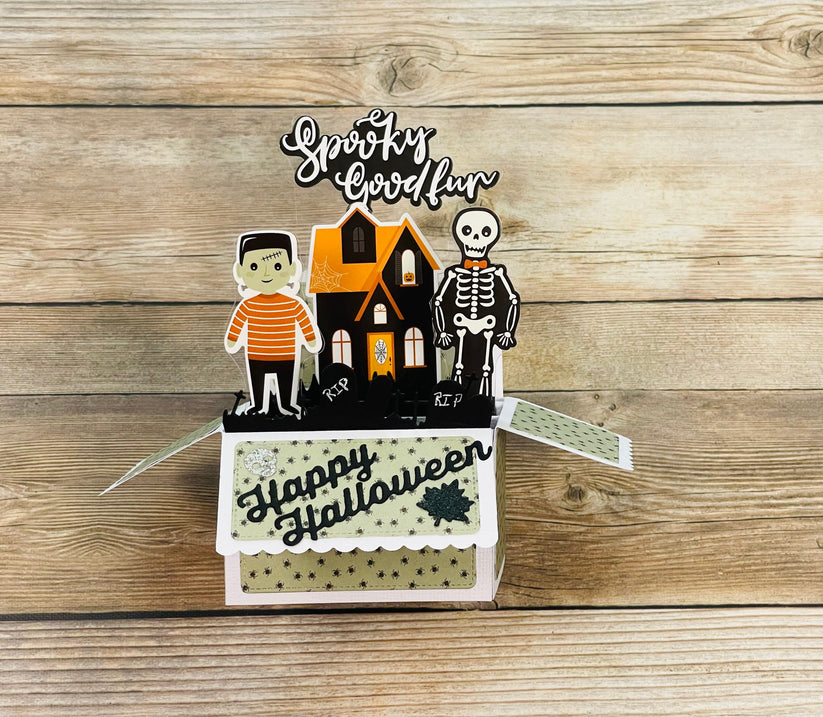 Halloween-themed box pop-up card a skeleton and Frankenstein monster, scary haunted house and cemetery. Message of Happy Halloween and spooky fun on a wooden surface