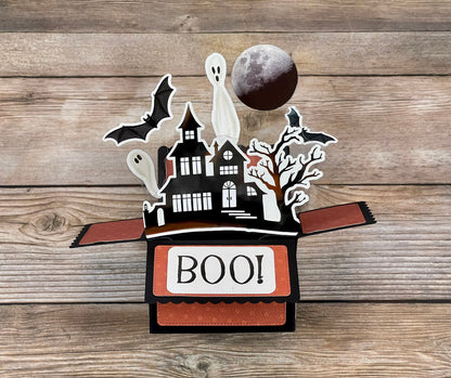 Halloween-themed pop-up box with ghosts, bats, and a moon on a wooden surface