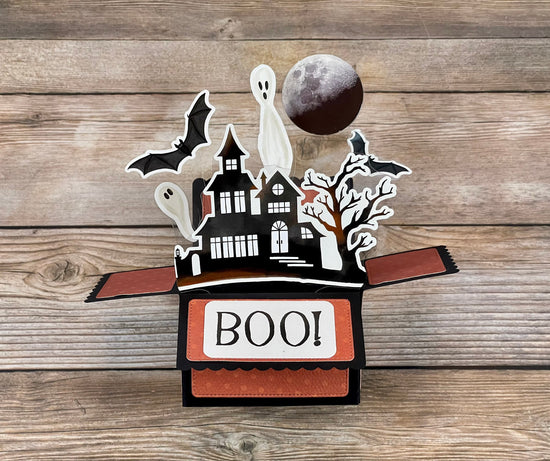 Halloween-themed pop-up box with ghosts, bats, and a moon on a wooden surface
