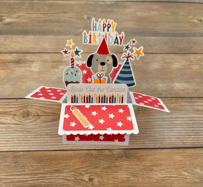 Pop-up birthday card with dog illustration, candles, and 'Happy Birthday' text on a wooden surface.