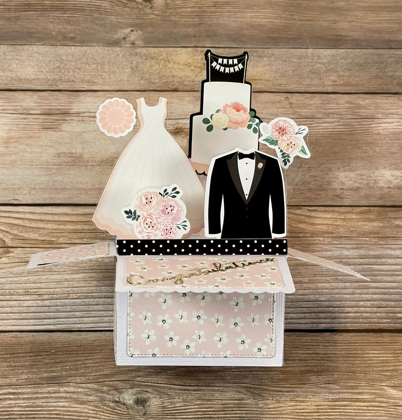 Card with wedding-themed cutouts of a dress, tuxedo, and cake on a wooden background