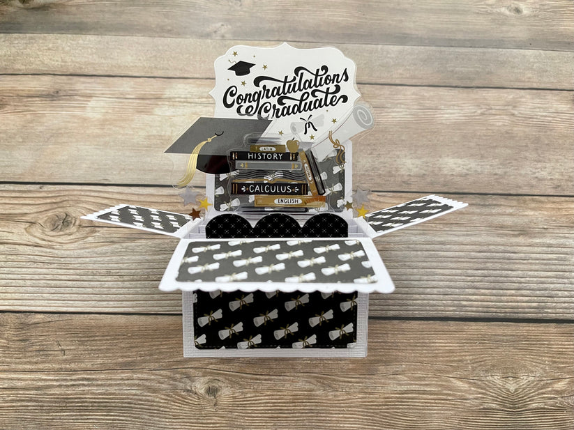 Graduation-themed pop-up card with books and mortarboard on a wooden surface