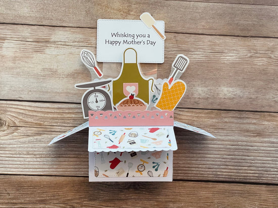 Pop-up card with kitchen-themed illustrations and a card inside on a wooden surface