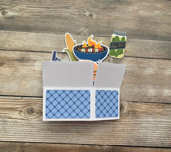 Card with cut-out graphics of a grill, corn, and beer on a wooden surface