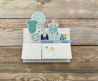Baby-themed card with illustrations of baby clothes and shoes on a wooden surface
