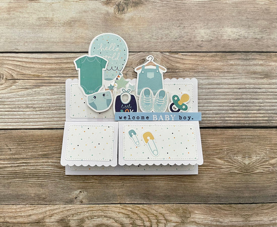Baby-themed card with illustrations of baby clothes and shoes on a wooden surface