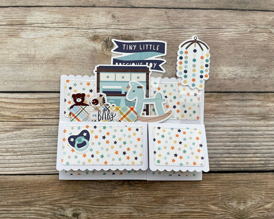 Baby-themed card with illustrations of a crib, baby bottle, and pacifier on a wooden surface.