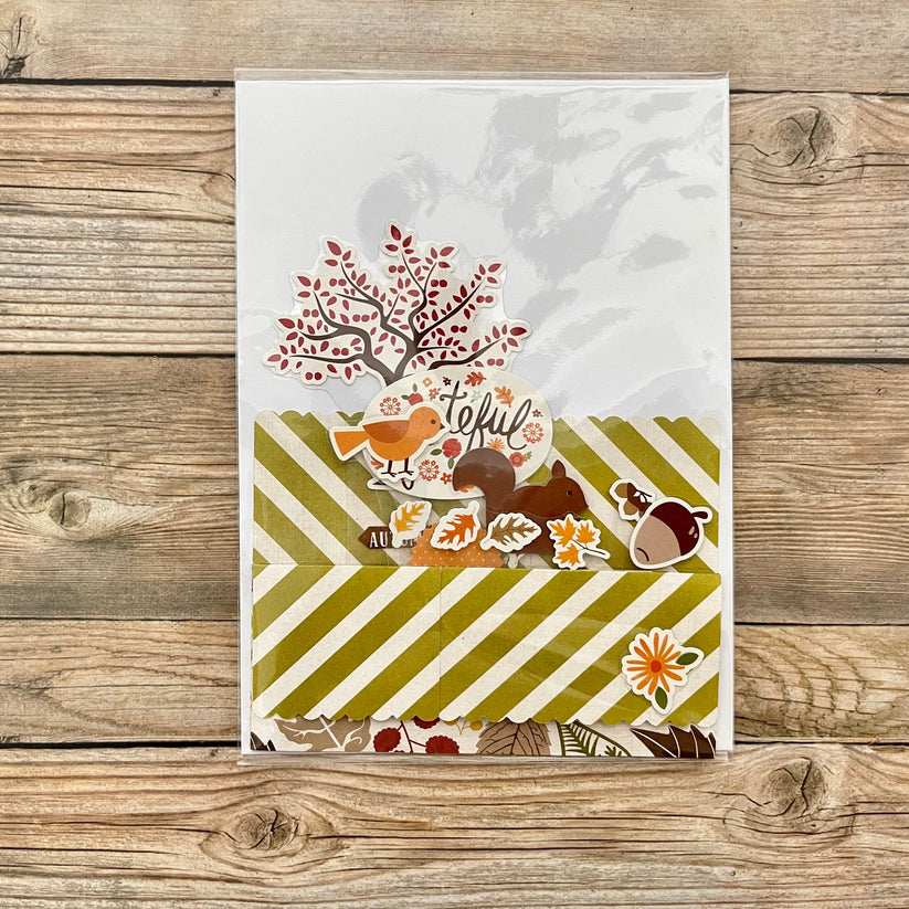 Autumn-themed card with trees, birds, and acorns on a wooden surface