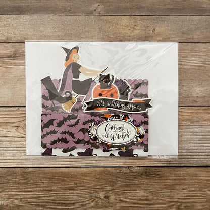 Halloween-themed card with a witch and cats on a wooden surface