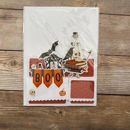 Halloween-themed card with skeleton, bat, and 'BOO' banner on a wooden surface