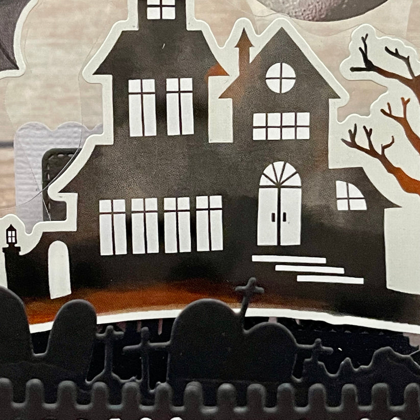 Halloween-themed cutout with a haunted house, trees, and a moon on a gray background