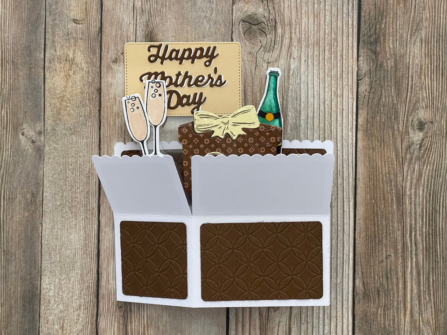 Card with Mother's Day message, champagne glasses, gift box, and bottle on a wooden background