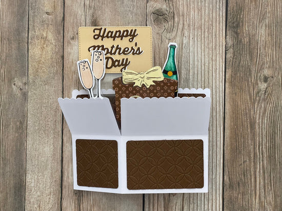 Card with Mother's Day message, champagne glasses, gift box, and bottle on a wooden background