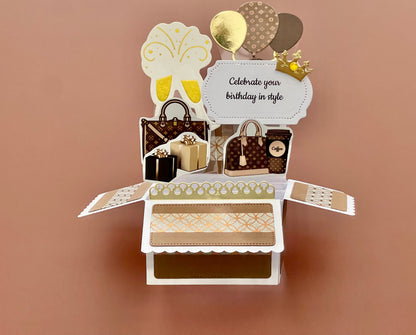Decorative birthday card with handbags, balloons, and a gift on a brown background