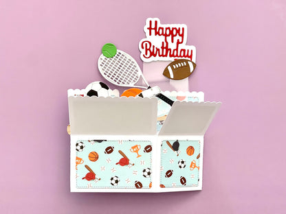 Open box with sports-themed items and a 'Happy Birthday' topper on a pink background
