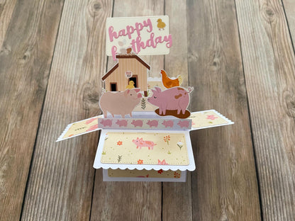 Pop-up birthday card with pigs, a house, and a duck on a wooden surface