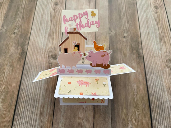 Pop-up birthday card with pigs, a house, and a duck on a wooden surface